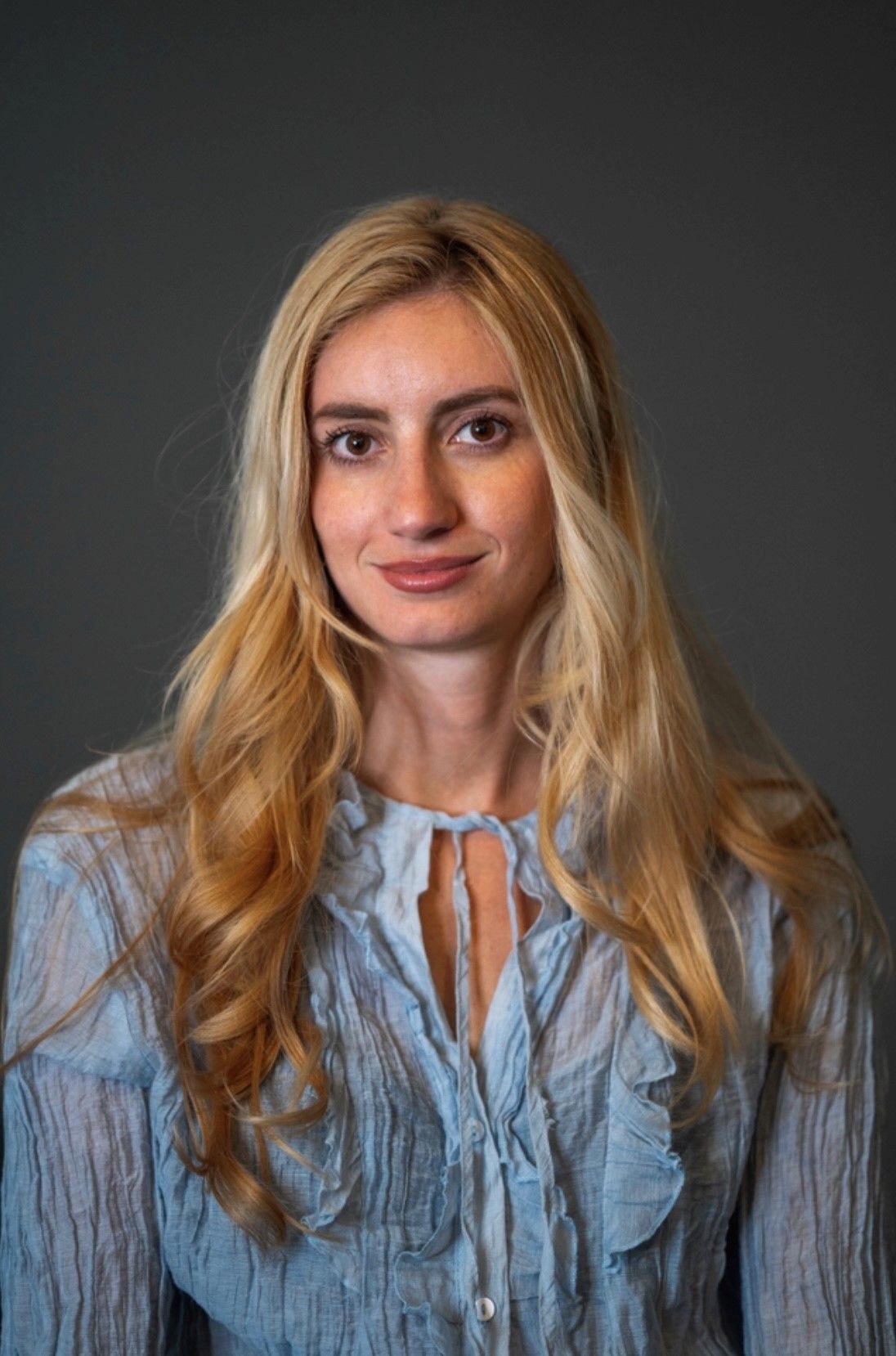 Woman with long blonde hair wearing a light blue blouse, posed against a plain dark background.